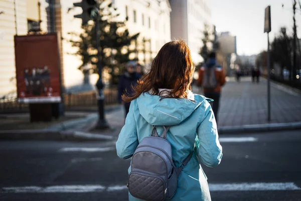 青いジャケットと小さなバックパックで散歩中の女の子の観光客は、道路を横切ります。後ろから見る. — ストック写真