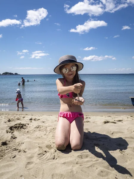 Vacations by the sea: a little girl in sunglasses and a hat pours her hands on the sand on a sea beach near the water.