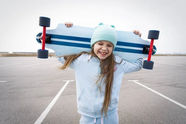 Een meisje in een blauwe hoed houdt een groot Longboard op haar schouders en lacht gelukkig met haar ogen gesloten. — Stockfoto