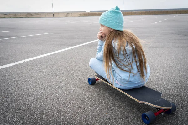 Een meisje in een blauwe muts en trui zit op een groot Longboard langs een groot asfalt terrein. — Stockfoto