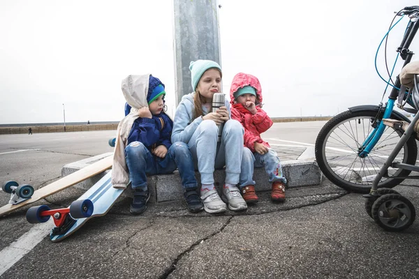 Drie kinderen fietste op een skateboard en een fiets en gingen zitten om te rusten op de stoeprand. — Stockfoto