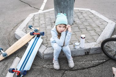 Mavi kazak ve şapkalı bir kız uzun tahtaya biniyordu ve bir termostan sıcak çay içmek için kaldırıma oturdu..