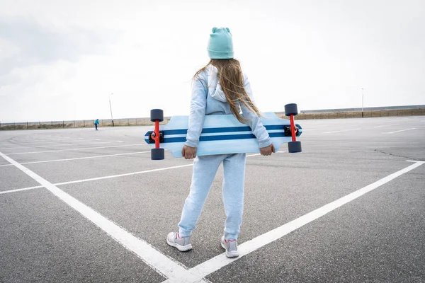 Een meisje in een hoed en een blauwe trui houdt een groot Longboard. — Stockfoto