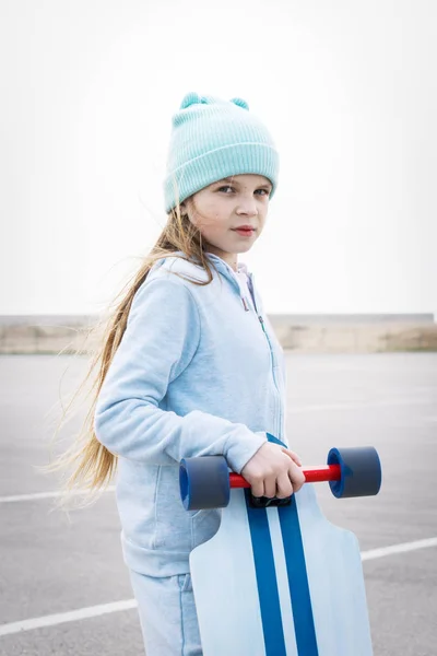 Een meisje in een hoed en een blauwe trui houdt een groot Longboard. — Stockfoto