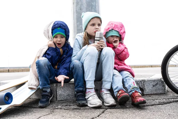 Drie kinderen fietste op een skateboard en een fiets en gingen zitten om te rusten op de stoeprand. — Stockfoto