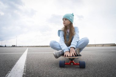 Mavi şapkalı ve kazakgiyen bir kız, büyük bir asfalt zemin boyunca büyük bir uzun tahtanın üzerinde oturuyor..