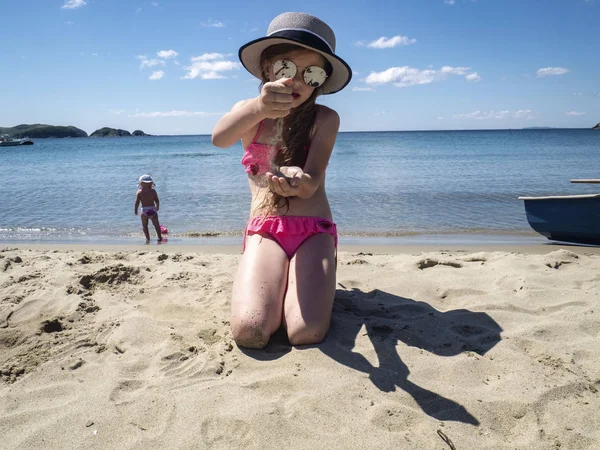 Vacations by the sea: a little girl in sunglasses and a hat pours her hands on the sand on a sea beach near the water.