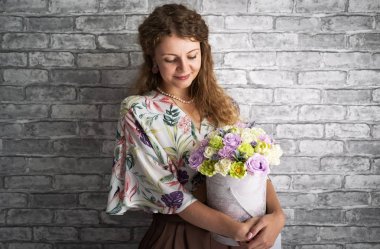 Sarışın kıvırcık saçlı bir kız tutar ve gri bir duvara karşı çiçeklerle büyük bir kutu kucaklar.