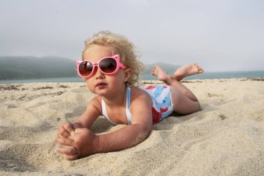 Küçük kıvırcık kız çocuk üç yaşında komik bir mayo denizde kum yatıyor. Büyük pembe güneş gözlüğü.