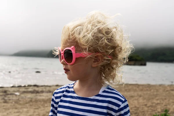Pequeña niña rizada en un chaleco se encuentra en la playa en gafas de sol y el viento voltea su cabello . — Foto de Stock