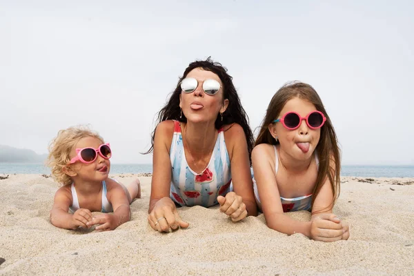Мама и две дочери в одинаковых купальниках и солнцезащитных очках лежат на песке и показывают язык. Младшая дочь с вьющимися волосами в розовых солнечных очках . — стоковое фото