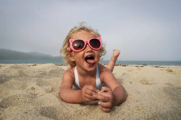 Niña rizada de tres años con un divertido traje de baño tirado en la arena junto al mar. Gafas de sol rosas grandes . — Foto de Stock