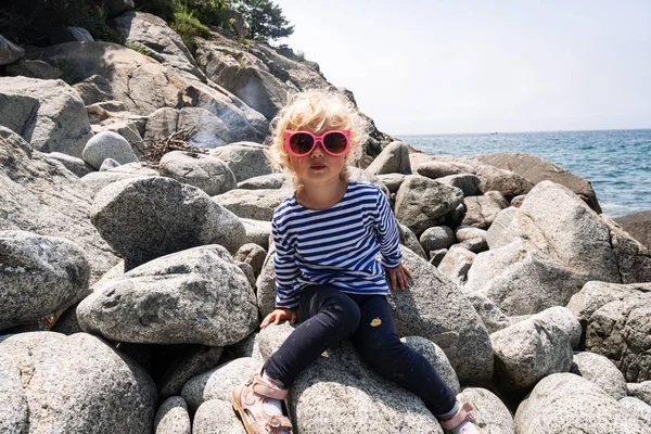 Pequeña niña rizada en un chaleco y gafas de sol de color rosa se sienta en enormes piedras junto al mar . — Foto de Stock