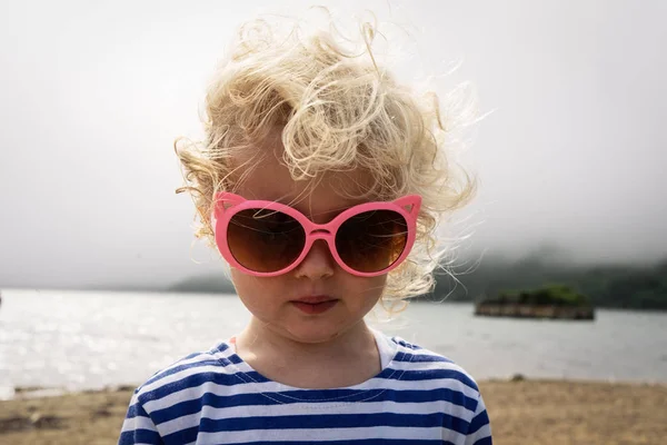 Kleine krullend meisje baby in een vest staat op het strand in zonnebril en de wind ruches haar haar. — Stockfoto