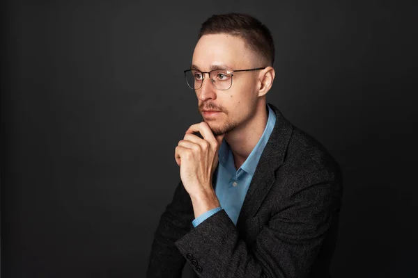 Interview: Studio portrait of a pensive young journalist guy with glasses with a beard on a dark background.