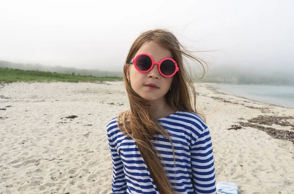 Meisje kind in zonnebril en een gestreepte vest op een mistige kust. Portret. — Stockfoto