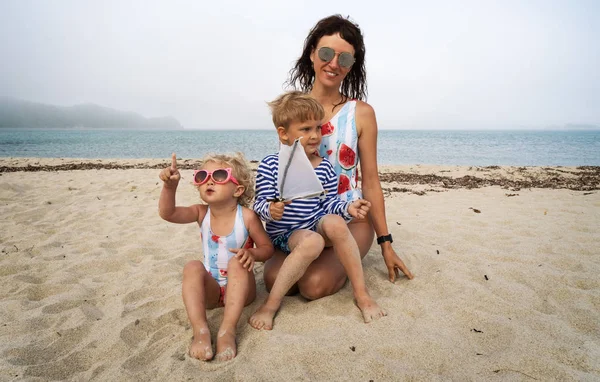 Мама с детьми дочь и сын в солнечных очках на берегу моря. Мальчик держит в руке импровизированную лодку . — стоковое фото