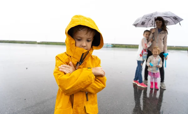 黄色いレインコートを着た小さな男の子が、家族から離れて雨の中に一人で立っている. — ストック写真