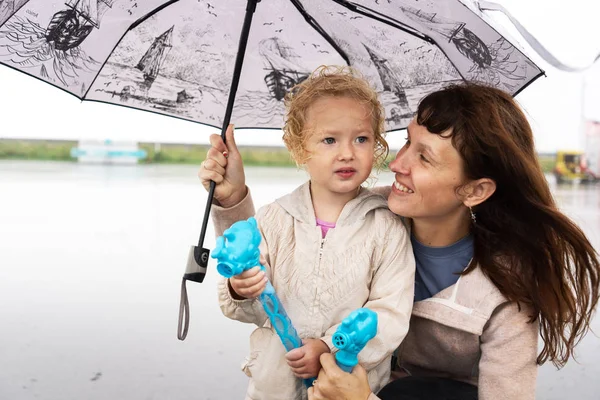 Mutter und kleine Tochter mit blonden Locken verstecken sich unter einem großen Regenschirm. — Stockfoto