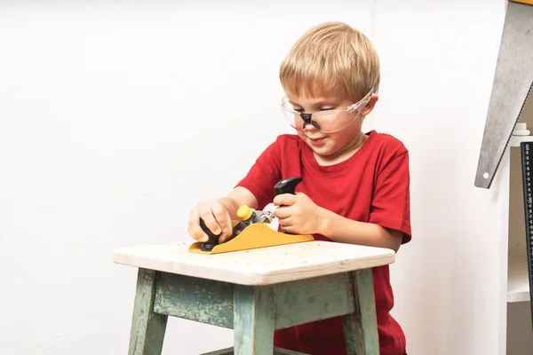 Um rapazinho planeia um banco velho com uma plaina. Treinamento nas habilidades de um carpinteiro e um marceneiro . — Fotografia de Stock