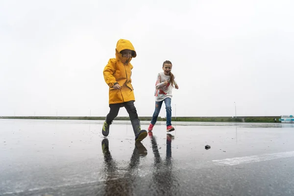 Egy kisfiú a sárga esőkabát ugrik át pocsolya az esőben. — Stock Fotó