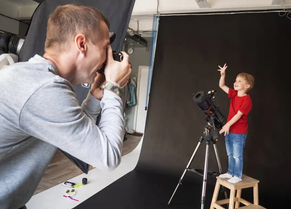 Un hombre toma un retrato de un niño con un telescopio en el estudio con luz artificial sobre un fondo negro . — Foto de Stock