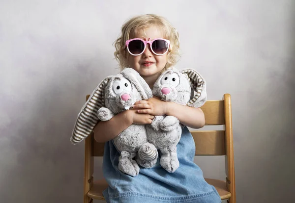 Niña en gafas de sol abraza dos conejos de felpa . — Foto de Stock