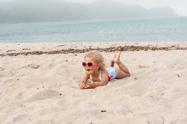 A little girl in sunglasses lies on the sand by the sea. — Stock Photo, Image