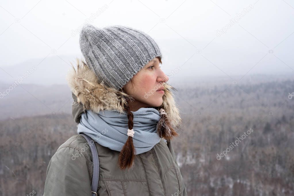 A girl in a winter hat and with pigtails stands on top of a mountain in the forest.