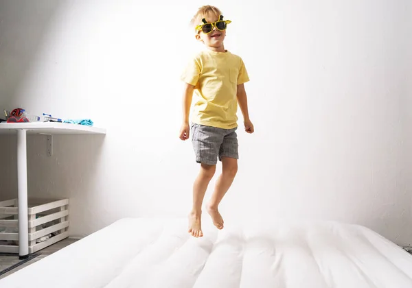 Ένα μικρό αγόρι με κίτρινα γυαλιά ηλίου και κίτρινη φούστα πηδάει σε ένα λευκό στρώμα. Πορτραίτο στούντιο σε λευκό φόντο. — Φωτογραφία Αρχείου