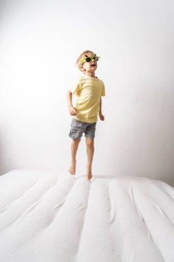A little boy in pink sunglasses and a yellow skirt jumps on a white mattress. Studio portrait on a white background.