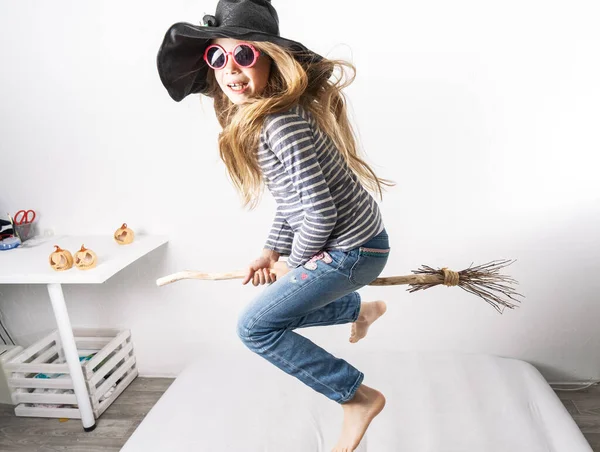 Una colegiala con un sombrero de bruja salta sobre un colchón y vuela sobre un palo de escoba. Retrato de estilo de vida infantil. —  Fotos de Stock