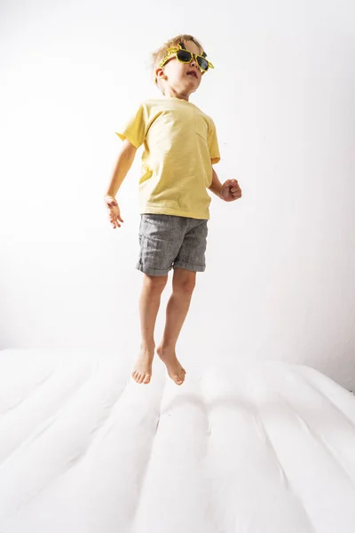 Un niño pequeño con gafas de sol amarillas y una falda amarilla salta sobre un colchón blanco. Retrato de estudio sobre fondo blanco . —  Fotos de Stock