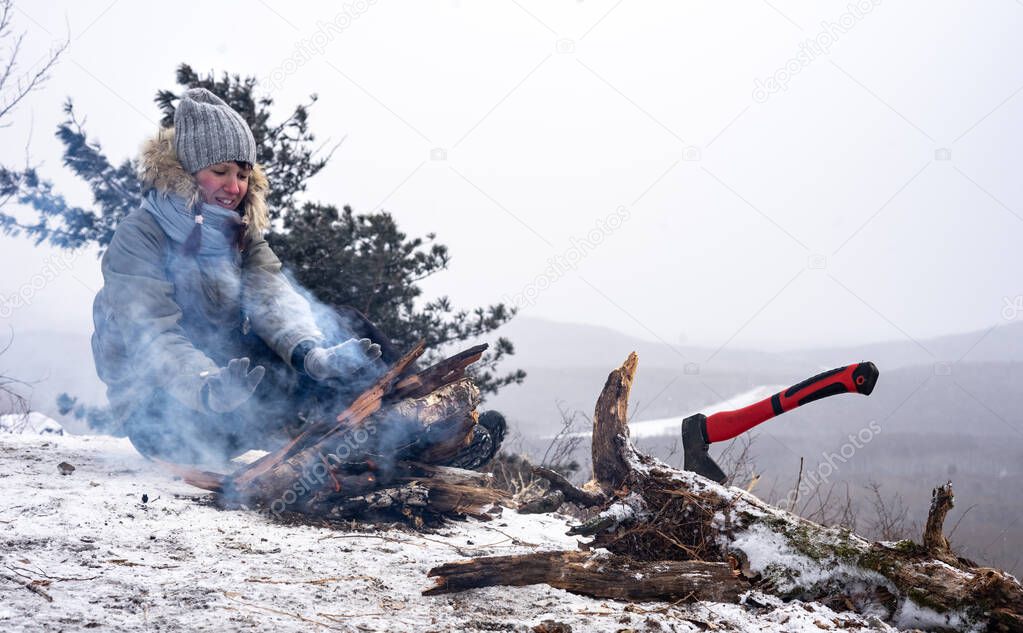 Girl hiker sits in the snow near a fire and basks.
