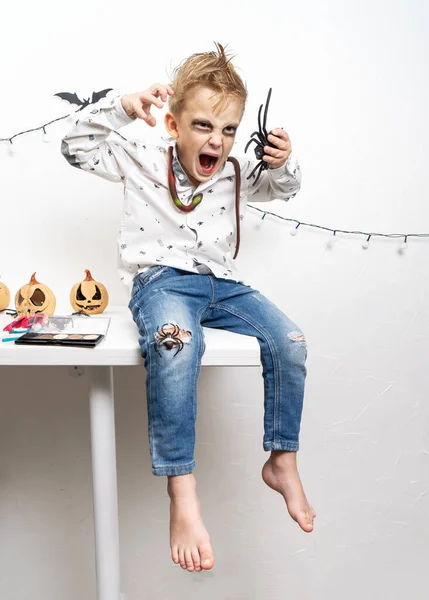 Un niño pequeño con un disfraz de zombi se sienta en una mesa y hace muecas. —  Fotos de Stock