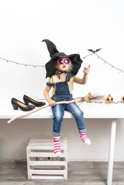 Feliz Halloween: Una niña se sienta en una mesa en un sombrero de bruja con una escoba. —  Fotos de Stock
