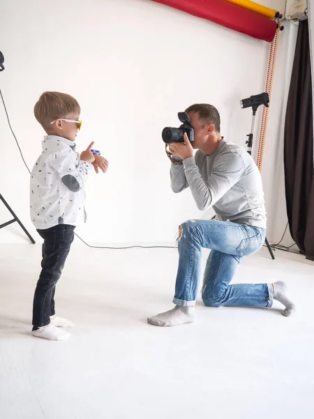 Fotógrafo hombre fotografías de un niño pequeño en una camisa blanca sobre un fondo blanco . — Foto de Stock