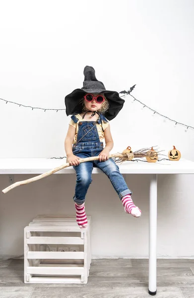 Feliz Halloween: Una niña se sienta en una mesa en un sombrero de bruja con una escoba. —  Fotos de Stock