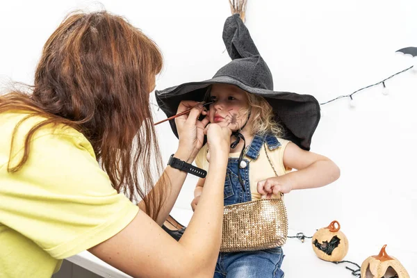 Mamá pone maquillaje en la cara de su hija en un disfraz de una pequeña bruja con una escoba y un sombrero . —  Fotos de Stock