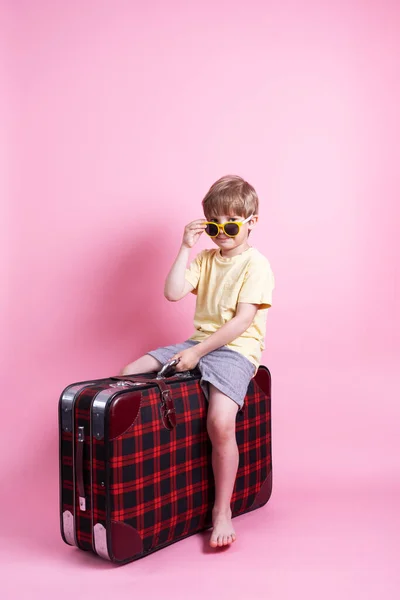 Viajar: Um menino de óculos de sol joga um turista e senta-se em uma grande mala vermelha . — Fotografia de Stock