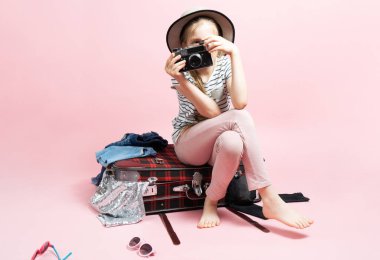 Seyahat: Bir kız öğrenci bir yolculuğa hazırlanıyor, büyük bir bavulun içinde oturuyor ve elinde büyük bir retoro kamerası tutuyor..