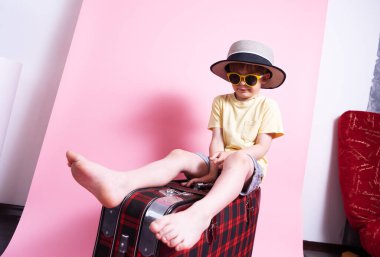 Seyahat: Hasır şapkalı ve güneş gözlüklü küçük bir çocuk turisti oynuyor ve büyük kırmızı bir bavula oturuyor.