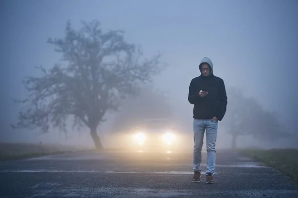 Joven Caminando Por Carretera Usando Teléfono Coche Acercándose Espesa Niebla —  Fotos de Stock