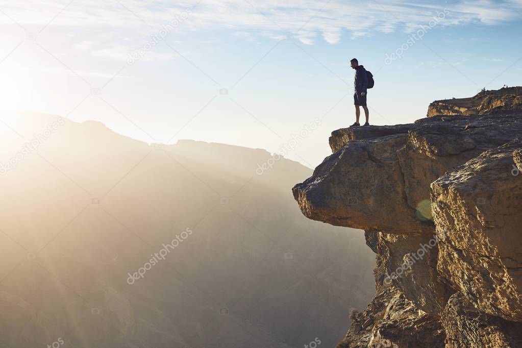 Young tourist with backpack standing on the edge of cliff at sunrise. Jebel Akhdar, Grand Canyon of Oman.  