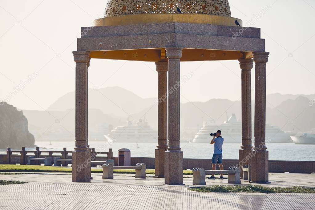 Young tourist photographing waterfront against harbor with modern passenger ships. Mutrah Corniche in Muscat, Sultanate of Oman.  