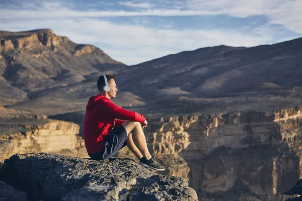 在山里放松 拿着耳机坐在悬崖边听音乐的年轻人 Jebel Akhdar 阿曼大峡谷 — 图库照片