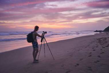 Young man photographing with tripod at beach after sunset. Photographer in blurred motion.   clipart
