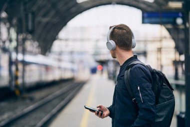 Kulaklıklı genç adam müzik dinliyor ve tren istasyonunda tren bekliyor.. 
