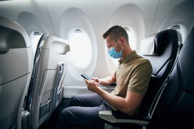 Yüz maskesi takan ve uçuş sırasında uçağın içinde telefon kullanan adam. Yeni normal, koronavirüs ve kişisel koruma temaları. 