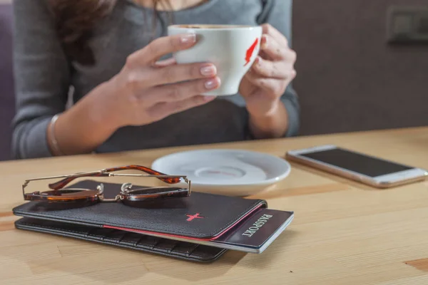 Female traveler drinking coffee at airport terminal, passport, ticket and smartphone on table